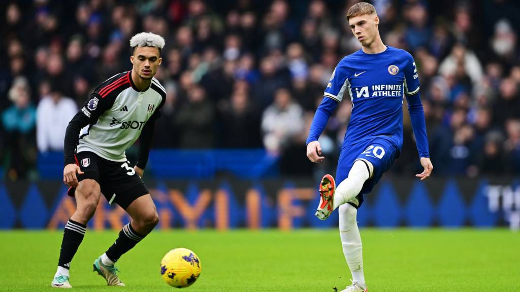 A Chelsea player in blue kit controls the ball while a Fulham player in black and white stripes watches closely during the Chelsea vs Fulham match.