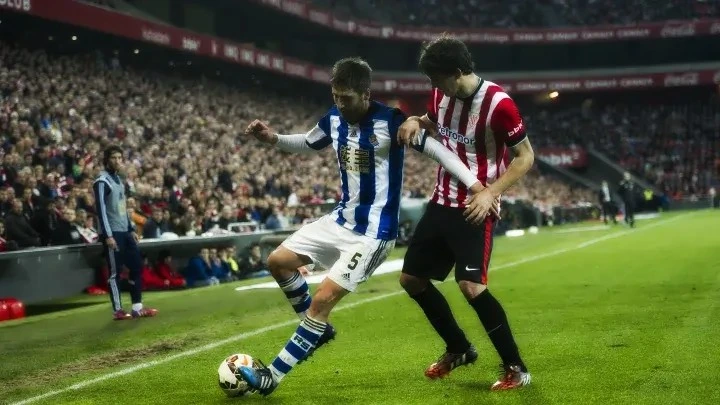 An Athletic Bilbao player in red and white stripes competes for the ball with a Real Sociedad player in blue and white stripes during a Bilbao vs Real Sociedad match.