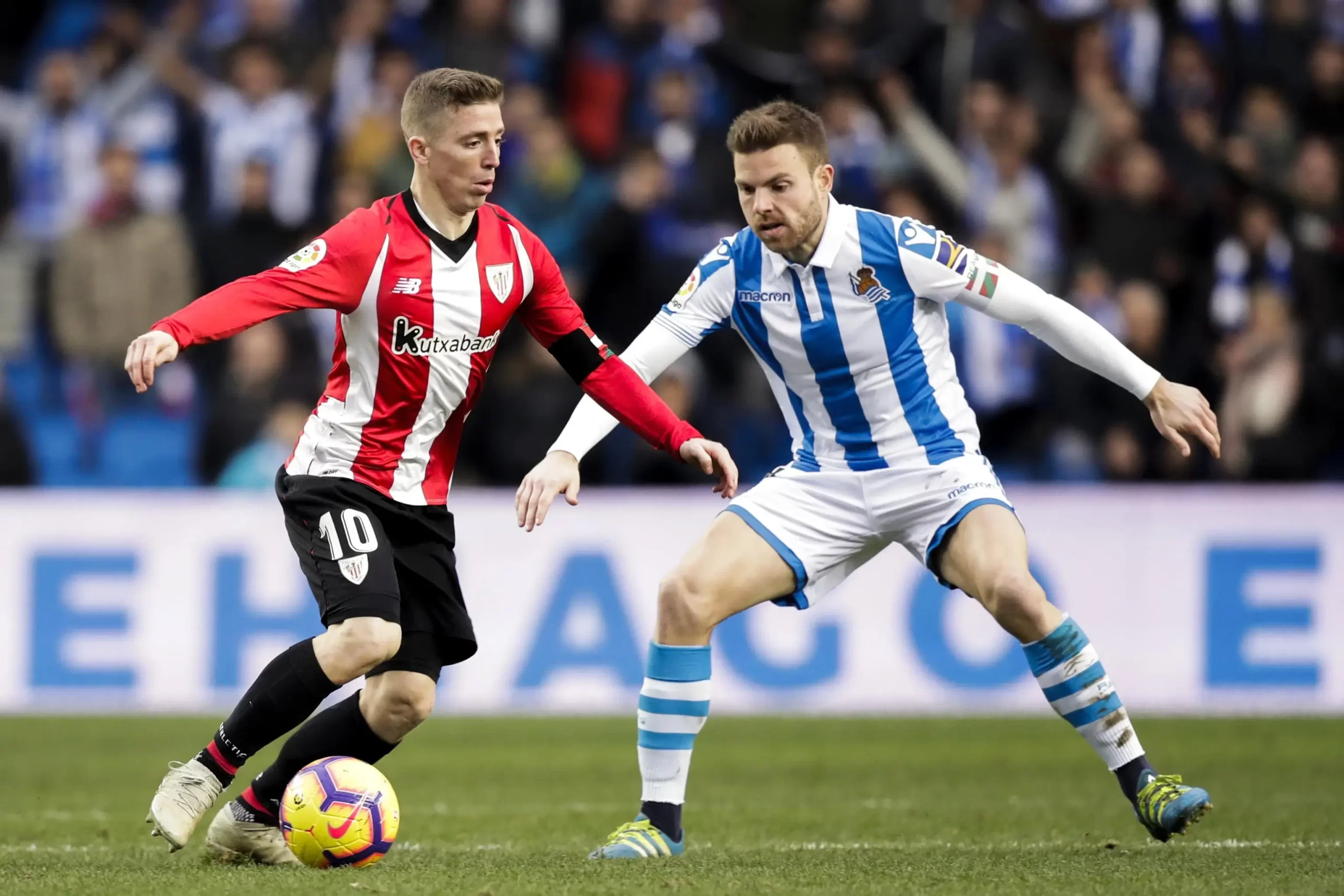 Two footballers, one from Athletic Bilbao in red and white and another from Real Sociedad in blue and white, vie for control of the ball on the pitch in a Bilbao vs Real Sociedad game.
